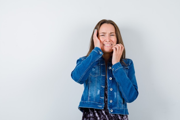 Señora joven con las manos cerca de la cara en blusa, chaqueta de mezclilla y mirada nostálgica. vista frontal.