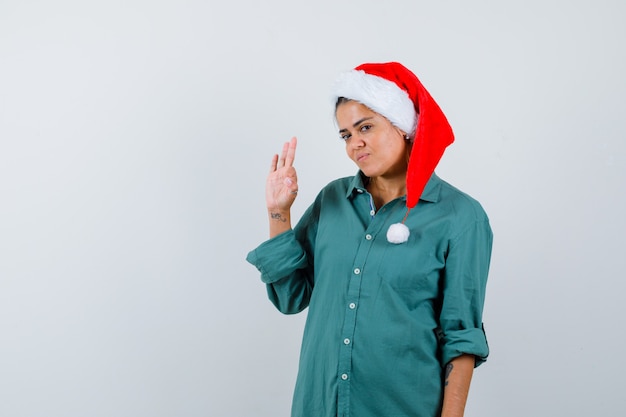 Señora joven que muestra el gesto aceptable en el sombrero de la Navidad, la camisa y que parece confiada. vista frontal.