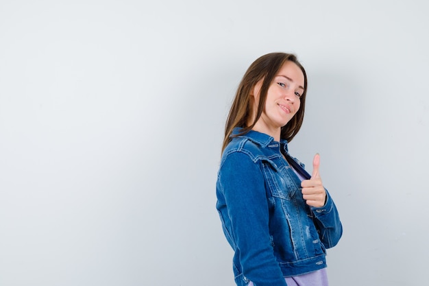 Señora joven que muestra el pulgar hacia arriba en camiseta, chaqueta y parece confiada.