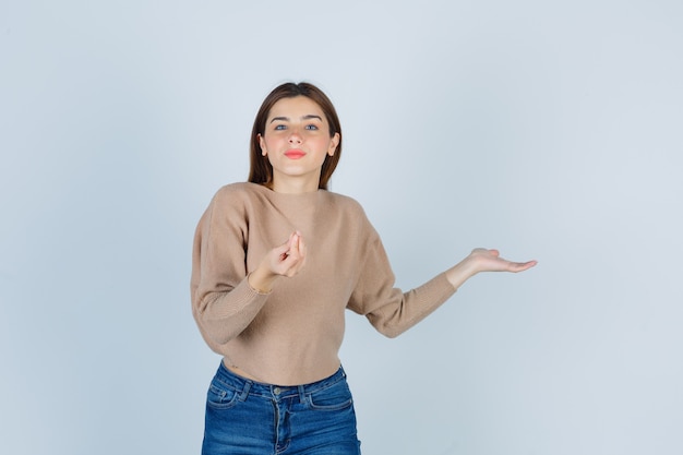 Señora joven en suéter beige, jeans mostrando un gesto delicioso y mirando complacido, vista frontal.