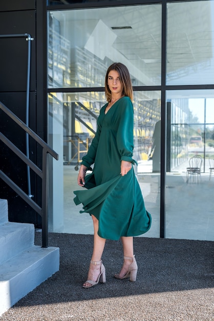 Señora joven en vestido verde de moda posign cerca del edificio de cristal, estilo de vida de verano