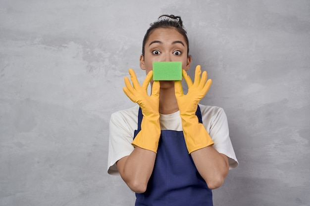 Señora de la limpieza divertida en uniforme y guantes de goma jugando con una esponja de cocina mirando a la cámara