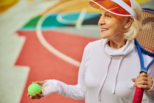 Señora mayor, saludable y alegre en una colorida plaza al aire libre y sosteniendo la raqueta de tenis con pelota