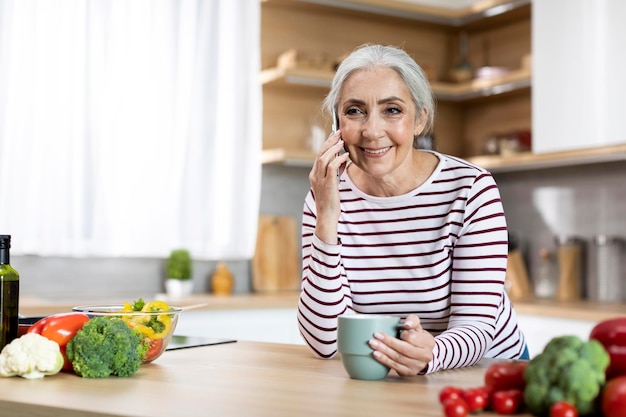 Señora mayor sonriente hablando por teléfono celular y bebiendo café en la cocina