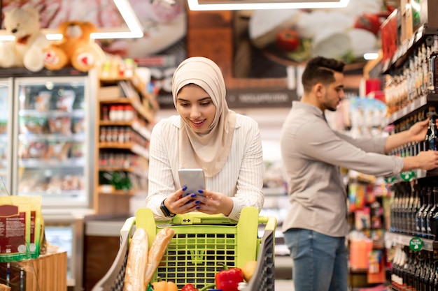 Señora del Medio Oriente comprando comestibles usando el teléfono caminando en el supermercado