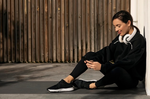 Señora milenaria caucásica sonriente en ropa deportiva y auriculares descansando sentada en el suelo escribiendo en el teléfono inteligente