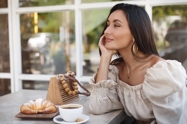 Señora de moda en un café, tomando café, apoyado en la mesa y disfrutando de la vista al aire libre.