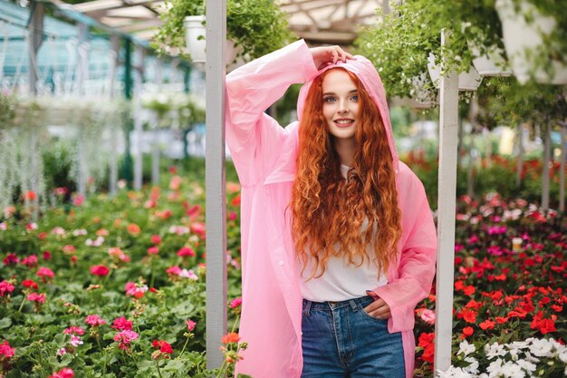 Señora muy sonriente con el pelo rizado pelirrojo de pie en un impermeable rosa y mirando soñadoramente a la cámara mientras pasa tiempo en el invernadero con hermosas flores en el fondo