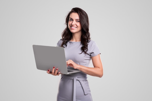 Señora de negocios adulta feliz en vestido elegante mirando a la cámara mientras trabaja en la computadora portátil contra el fondo gris