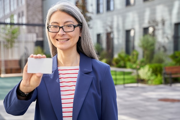 Foto señora de negocios madura sosteniendo una tarjeta de plástico en blanco y sonriendo