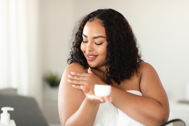 Foto señora negra de talla grande que aplica crema corporal en el hombro después de la ducha por la mañana haciendo un procedimiento de belleza