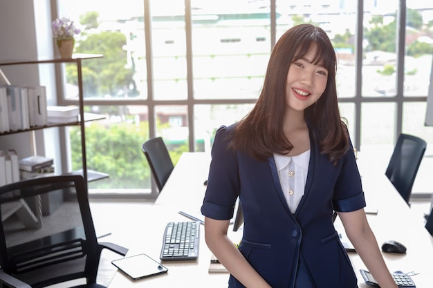 La señora de la oficina es hermosa, cabello largo, negro y feliz asiática con un traje azul oscuro en la oficina moderna con un escritorio y grandes ventanales que difuminan el fondo.