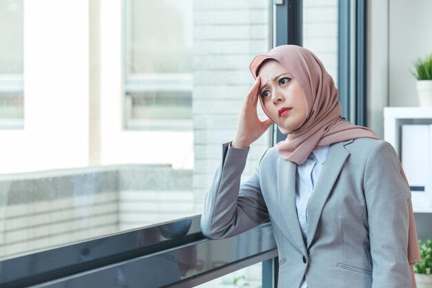 Señora de la oficina musulmana descansando junto a la ventana y con dolor de cabeza debido al cliente molesto. Concepto de trabajo negativo. mujer triste que trabaja en el trabajo de oficina.