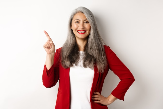 Señora de la oficina senior feliz que señala el dedo en la esquina superior izquierda, mostrando el logotipo de la empresa y sonriendo a la cámara, de pie sobre fondo blanco. Copia espacio