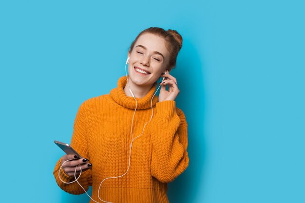 Señora pelirroja con pecas escuchando música con teléfono y auriculares está planteando en una pared azul