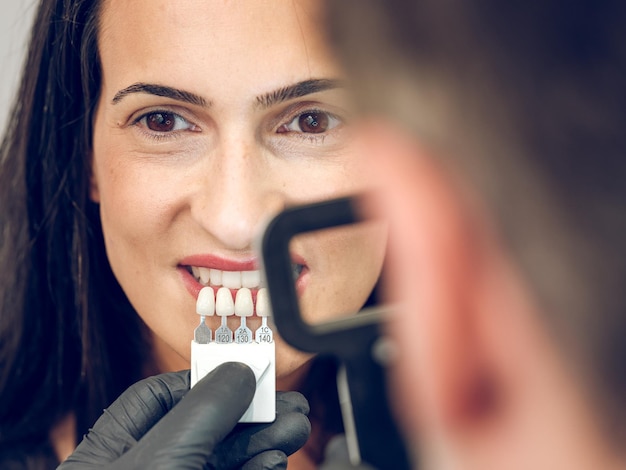 Señora de pelo oscuro mirando a la cámara mientras el dentista borrosa selecciona el tono de los dientes con lupa y escala de sombra
