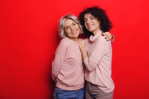 Señora de pelo rizado con su amiga rubia abrazando alegremente sobre un fondo rojo.