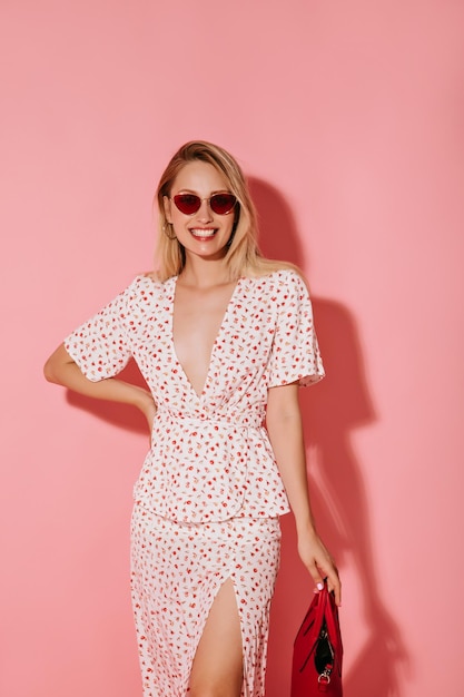 Señora positiva con cabello rubio en vestido ligero de moda y gafas de sol rojas sonriendo y sosteniendo un bolso rojo en un contexto aislado