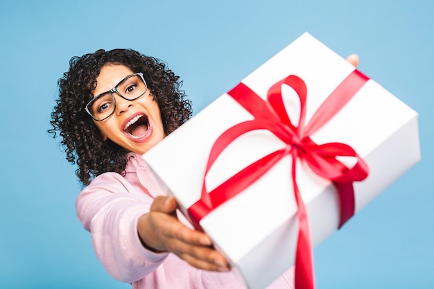 Señora rizada afroamericana feliz en la risa ocasional mientras sostiene la caja de regalo presente aislada sobre fondo azul.