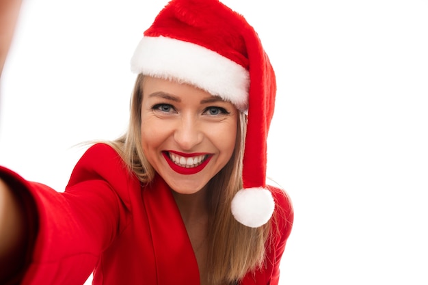 Señora rubia alegre en traje rojo y sombrero de Santa sosteniendo su mano mientras toma un selfie. Concepto de vacaciones