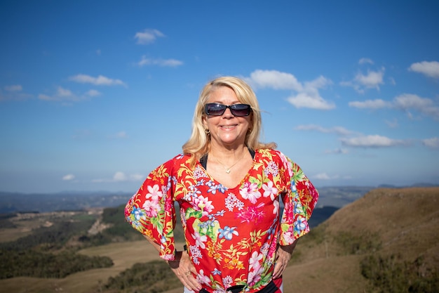 Señora rubia con blusa estampada y gafas de sol sonriendo en el hermoso paisaje de fondo