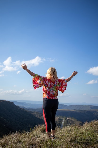 Señora rubia en blusa estampada y pantalón negro con los brazos extendidos mirando el paisaje