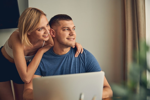 Señora rubia delgada disfrutando de un rato agradable mientras abraza a un hombre morena que trabaja en línea en la computadora