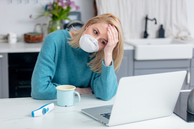 Señora rubia en máscara con taza de bebida, termómetro y computadora portátil en la cocina