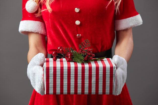 La señora Santa Claus vestida con la túnica roja y guantes blancos sostiene el regalo de Navidad en sus manos sobre el fondo gris. .