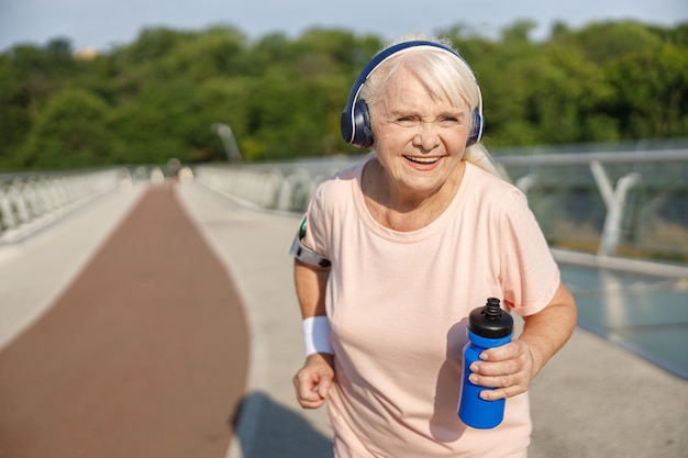 Señora senior positiva con auriculares corre a lo largo del puente en verano