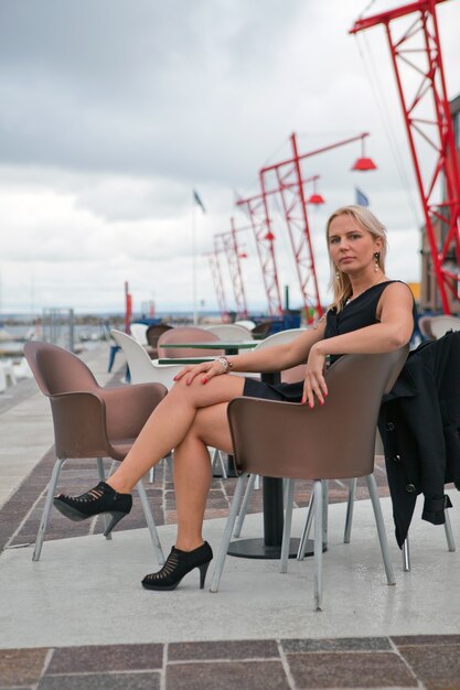 Señora sentada en la silla en la calle. Retrato de joven mujer bonita rubia sentada en el café de la calle sonriendo