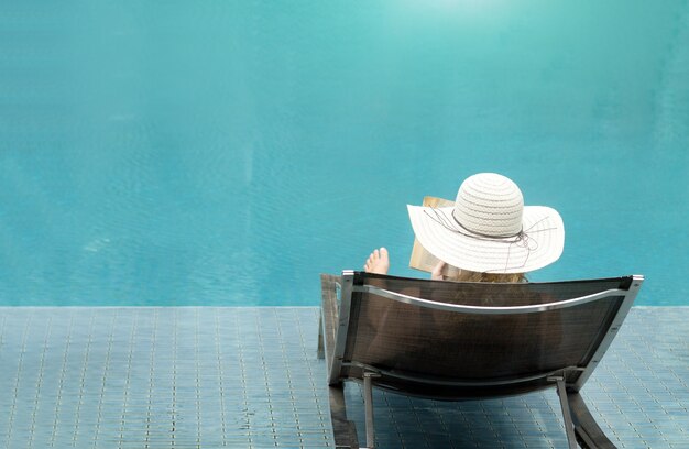 Señora con sombrero sentado y leer el libro junto a la piscina