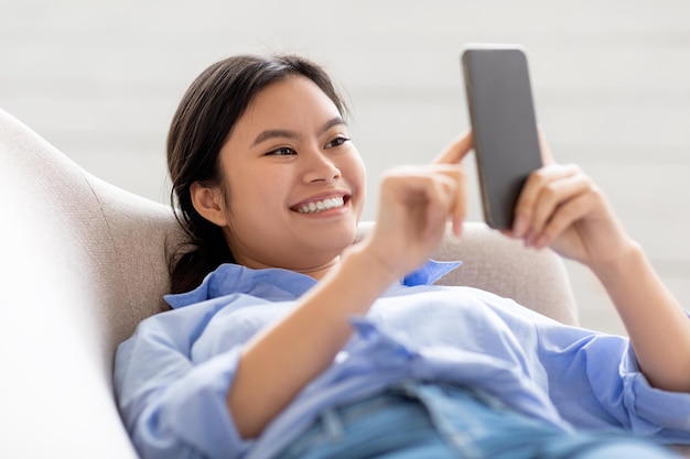 Señora sonriente recostada en el sofá usando el primer plano del teléfono celular