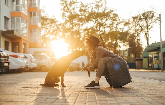 La señora sonriente se sienta con el perro atado contra el fondo de la puesta de sol y habla sobre el concepto de mascotas