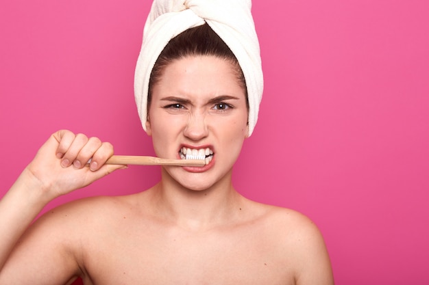 Señora sosteniendo su equipo dental en una mano, cepillándose los dientes cuidadosamente, cubriéndose el cabello con una toalla blanca