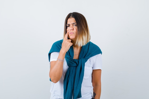 Señora con suéter atado apuntando a su párpado en camiseta blanca y mirando triste, vista frontal.