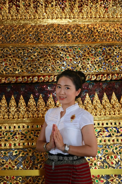 Foto señora tailandesa en traje tailandés pone las palmas de las manos juntas en saludo en la pared de mosaico de vidrio tailandés