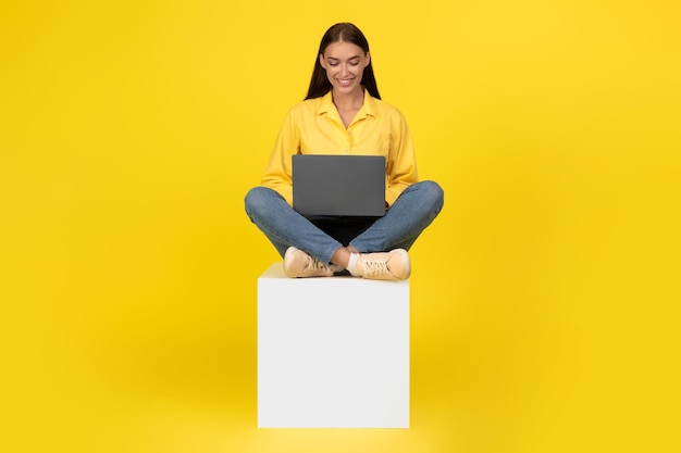 Señora trabajando en equipo portátil sonriendo sentada sobre fondo amarillo