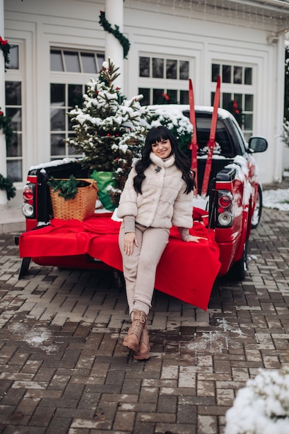 Señora vistiendo un abrigo de piel corto descansando en el borde de la carrocería de un camión rojo con árboles de Navidad y esquís.