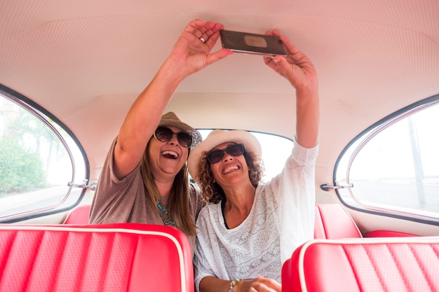 Señoras alegres caucásicas y selfie con moderno teléfono inteligente dentro de un viejo coche vintage rojo