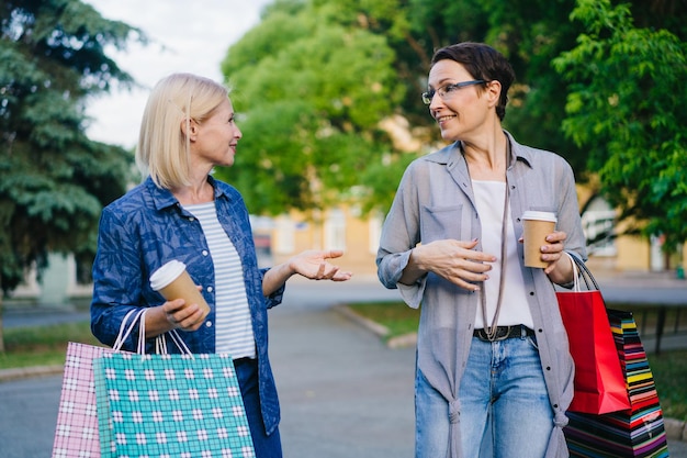 Señoras alegres hablando caminando en el parque con bolsas de compras disfrutando el fin de semana