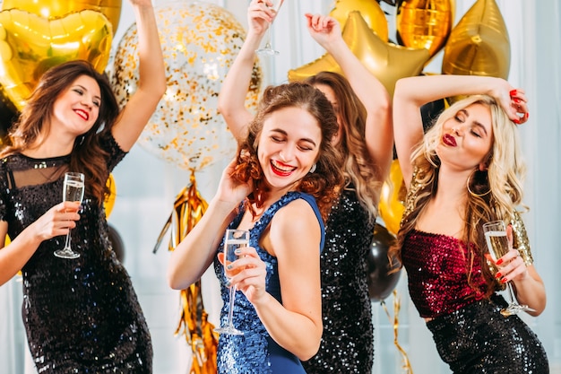 Señoras celebrando su graduación universitaria en casa. Chicas bailando con champagne en habitación decorada con globos.