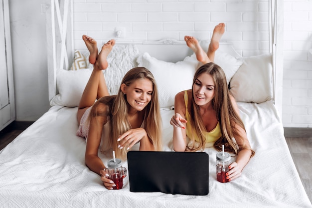 Señoras felices están acostadas en la cama relajándose en un carril de bebidas y buscando información en Internet