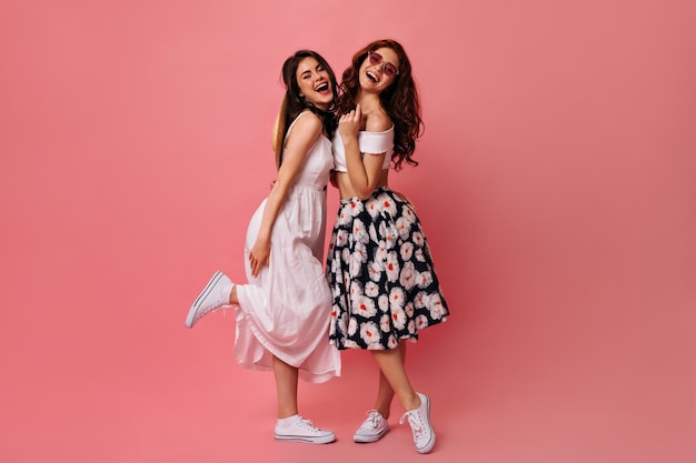 Señoras rizadas en vestidos midi posan sobre fondo rosa Chicas encantadoras con maquillaje brillante abrazando sonriendo y divirtiéndose en aislado