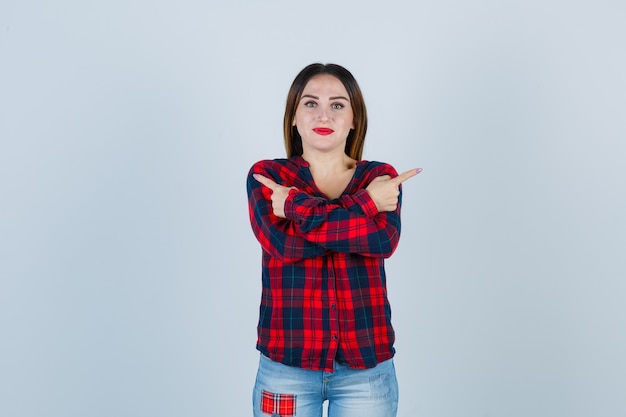 Señorita apuntando hacia los lados izquierdo y derecho en camisa a cuadros, jeans y luciendo alegre. vista frontal.