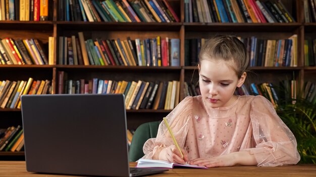 Señorita en blusa rosa mira la pantalla del portátil y escribe en el cuaderno sentado en la mesa de madera contra libros de colores en los estantes de la biblioteca