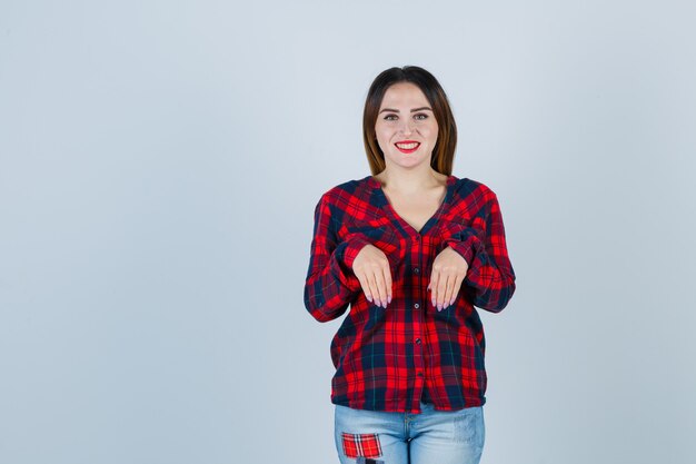 Señorita en camisa a cuadros, jeans manteniendo las manos sobre el pecho como palmas y mirando positivo, vista frontal.