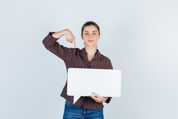 Señorita en camisa, jeans apuntando hacia abajo, manteniendo un cartel de papel y luciendo confiada, vista frontal.