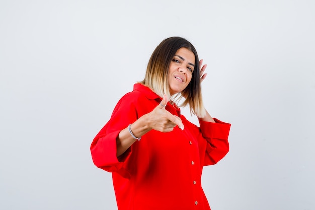 Señorita en camisa roja de gran tamaño apuntando a un lado y mirando alegre, vista frontal.