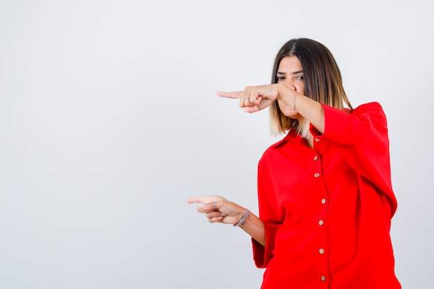 Señorita en camisa roja de gran tamaño apuntando a un lado y mirando complacido, vista frontal.